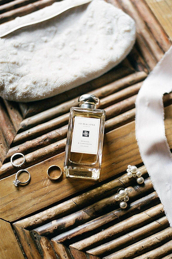 Wedding rings and engagement ring close up arranged with perfume bottle, pearl earrings, clutch purse, and ribbon on a wooden tray