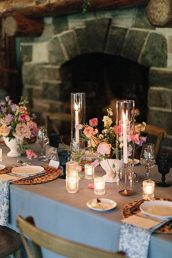Reception tablescape with a candlelit wedding table, floral centerpieces and taper candles in glass hurricanes set before a stone fireplace mantel