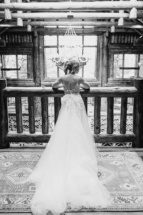 Bridal portrait of a bride in a backless wedding dress with lace bodice, leaning on a railing inside a rustic log cabin by large windows