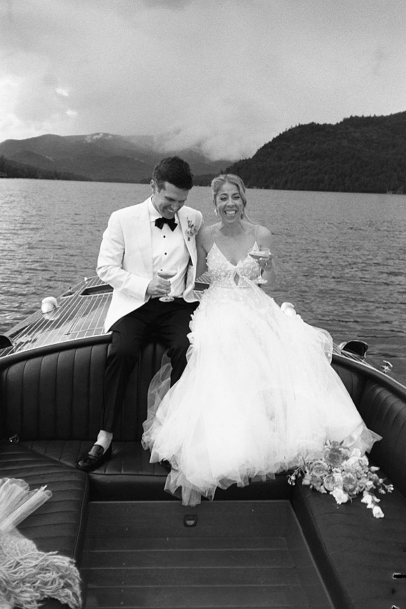 Couple portrait of newlyweds on boat, toasting with cocktail glasses as bride holds bouquet, with mountains and lake behind them