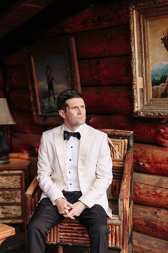 Groom portrait in a white tuxedo jacket with black bow tie, seated on a rustic wooden chair in a log cabin room with framed artwork