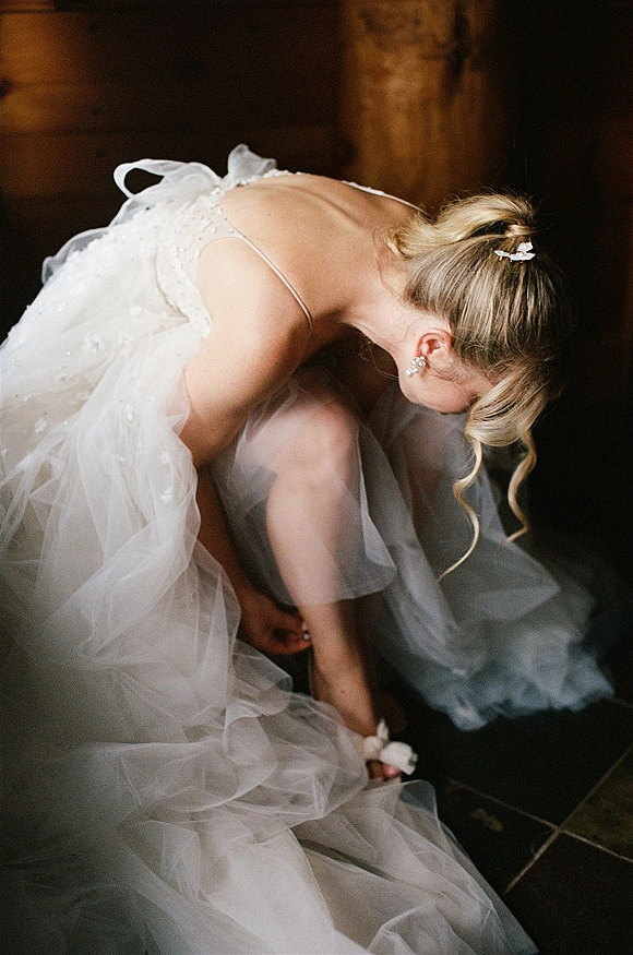 Bride getting ready as she puts on shoes, adjusting tulle wedding dress with spaghetti straps in a dark interior against a wood wall