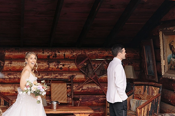 First look moment as bride approaches groom, bouquet and veil flowing in a rustic log cabin interior with wood walls and snowshoes