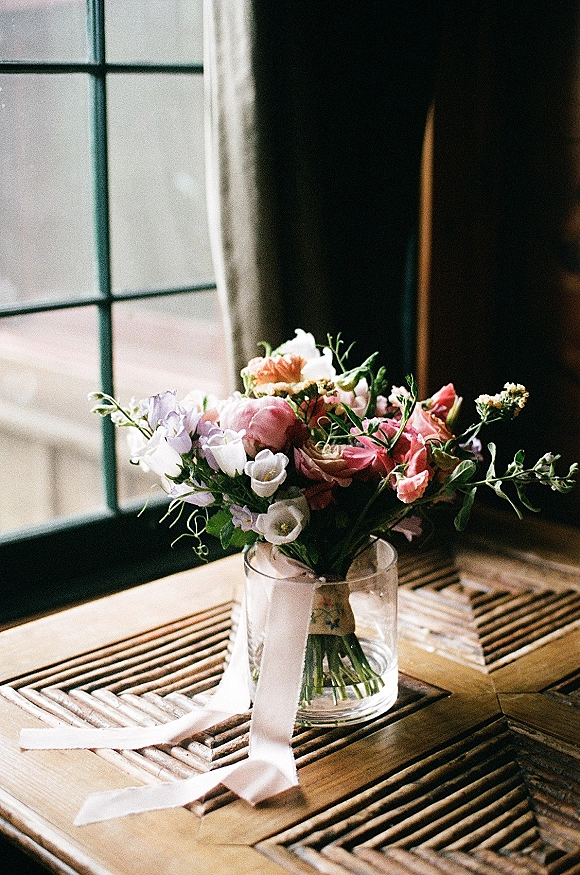 Bridal bouquet of mixed pastel flowers and greenery with ribbon in a glass vase on a wood table by window panes and curtains