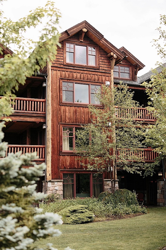 Rustic lodge exterior with timber beams and wood siding, large windows and stone foundation, framed by trees and lawn under sky