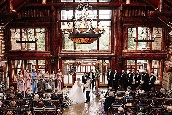Wedding ceremony with bride and groom at altar beneath floral arch, wedding party lined up in a log cabin with antler chandelier and forest windows