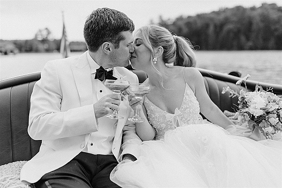 Wedding kiss portrait of bride and groom kissing while toasting champagne in a vintage convertible, lake and shoreline behind them