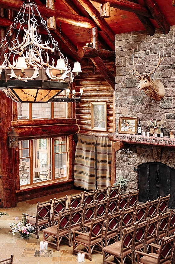 Ceremony setup with indoor ceremony seating, wood crossback chairs and candle-lit aisle facing a stone fireplace in a log cabin interior