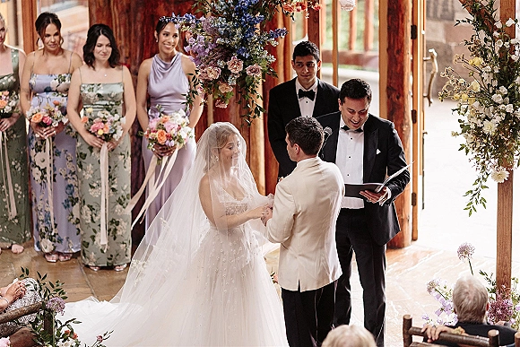 Wedding vows as bride in veil and groom in ivory jacket stand at floral ceremony arch inside rustic wood venue with guests seated behind