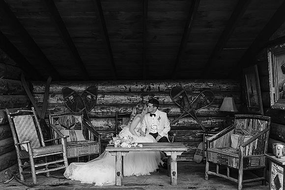 Wedding kiss portrait of bride and groom kissing on a bench in a log cabin lounge, with her dress train and bouquet by a wooden table