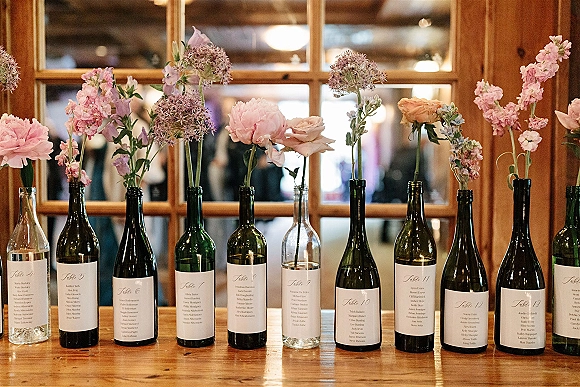Wedding escort display with wine bottle escort cards on a wood table, each bottle labeled and topped with pink flowers and peach roses indoors