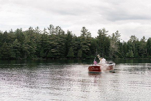 Wedding boat getaway as newlyweds wave from a wooden motorboat, bride in strapless dress raising bouquet, groom in white suit on a lake by forest shore