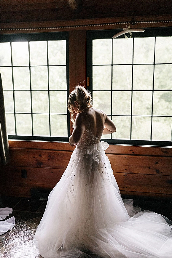 Bridal portrait of a bride by window in a lace bodice and tulle train, backless gown with bow sash, in a rustic cabin interior.