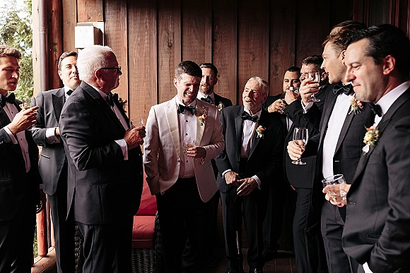Groomsmen portraits of the groom with groomsmen in tuxedos and bow ties, holding whiskey glasses by a rustic wood wall porch with trees behind
