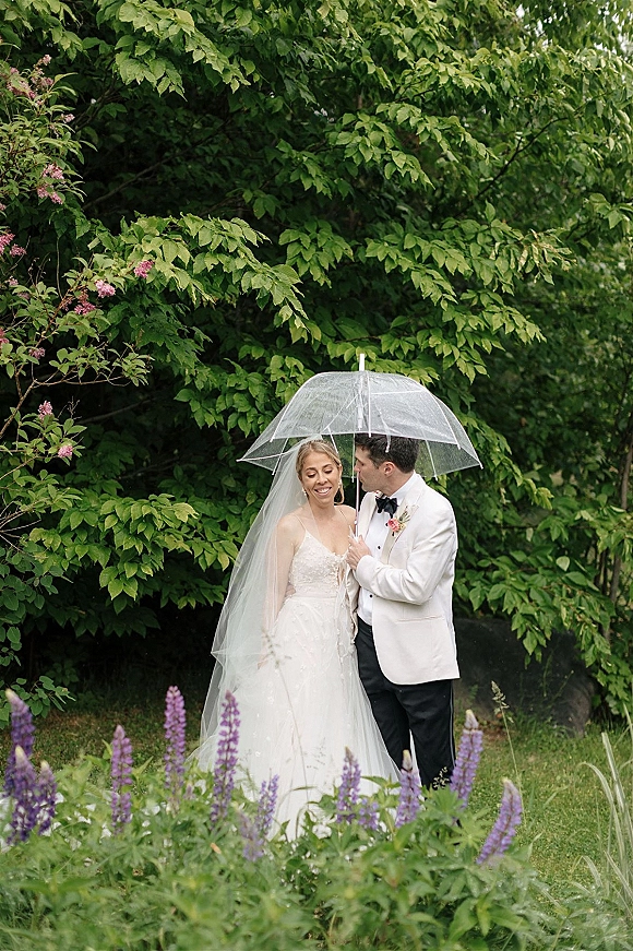 Wedding couple portrait of bride and groom under umbrella, her long veil and strapless dress beside his white tuxedo in a lush garden with purple flowers