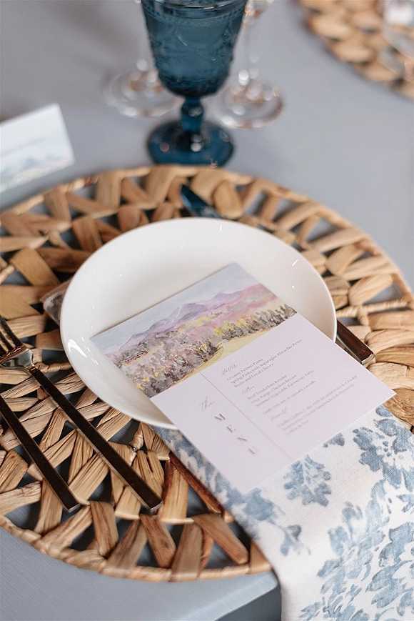 Wedding place setting with a wedding menu card on a white plate atop a woven rattan charger, blue goblet, gold flatware, gray tablecloth