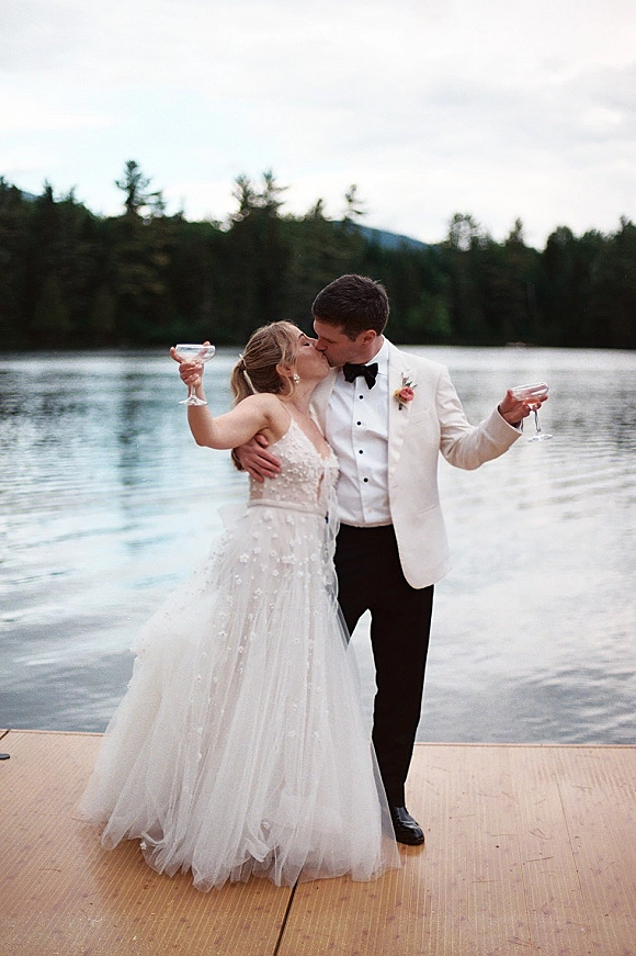 Wedding kiss as bride and groom toast with champagne coupe glasses on a dock, lake and mountains behind, tuxedo and gown visible