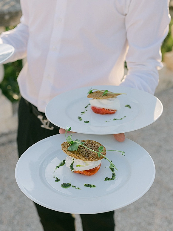 Wedding plated appetizer with microgreens garnish and herb sauce drizzle on a white plate, held by a server along an outdoor walkway with greenery