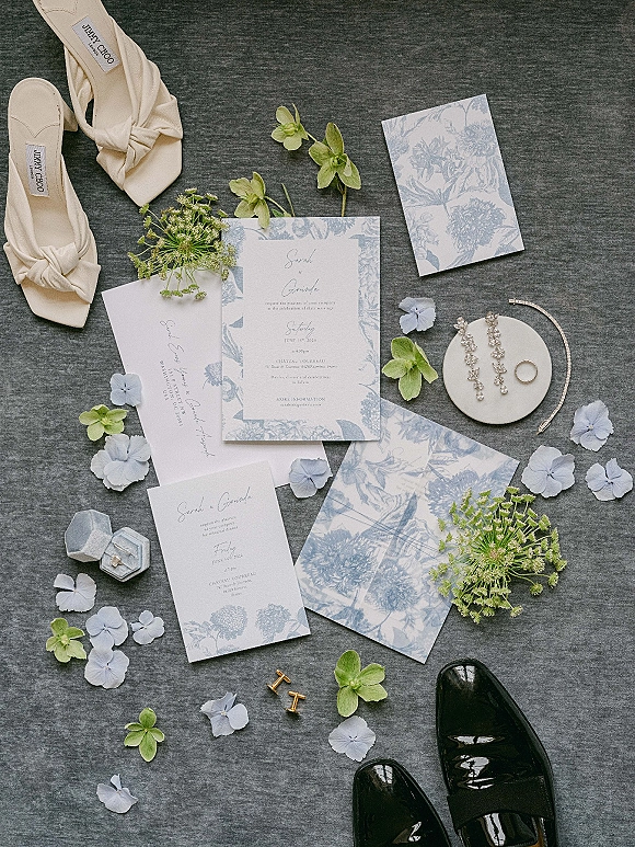 Invitation suite with blue floral wedding invitations, envelopes, wax seal, rings, earrings, cufflinks and shoes styled on gray fabric with petals
