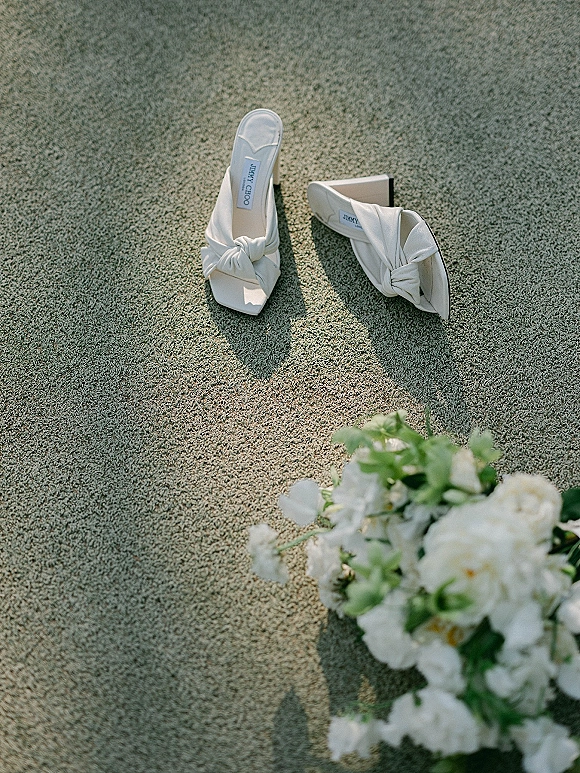 Wedding shoes in a bridal shoes flat lay, white heeled mules with bow detail beside a bouquet of white flowers and greenery on textured carpet