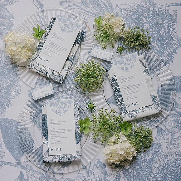 Wedding tablescape with a blue and white tablescape featuring clear glass plates, printed menu cards, toile linens, and hydrangea blooms