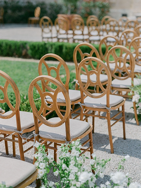 Ceremony seating with outdoor ceremony chairs in natural wood, lined along a gravel aisle with white flowers and greenery on a garden lawn