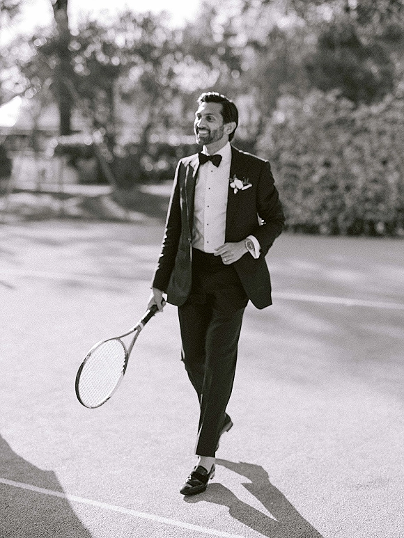Groom portrait in tuxedo with bow tie and boutonniere, holding a tennis racket on an outdoor tennis court with trees behind him