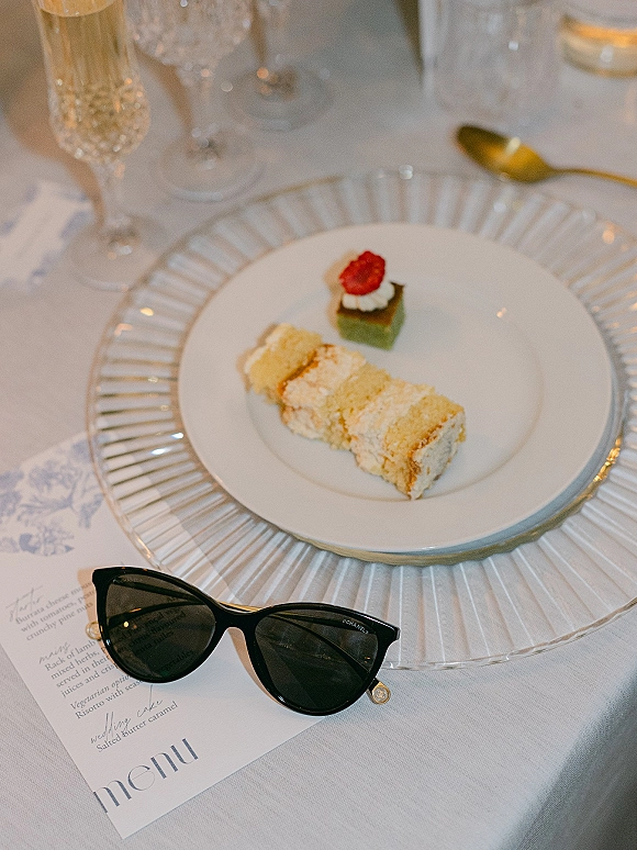 Wedding dessert plate with cake slice and petit four, raspberry garnish on white plate with gold spoon, menu card, and champagne flute on white linen