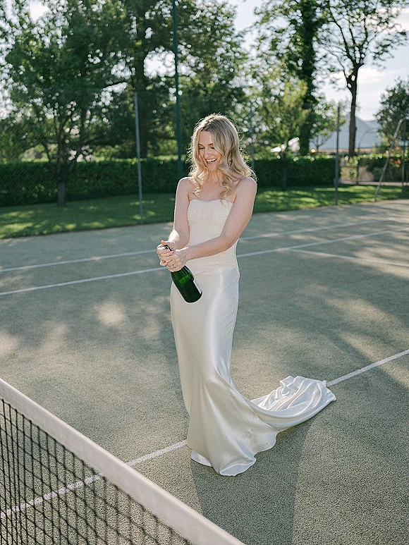 Bride portrait popping champagne in a strapless satin wedding dress on a tennis court, bottle spraying beside the net and greenery