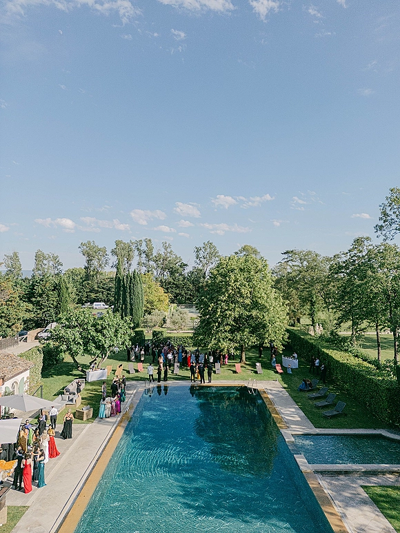 Outdoor wedding reception set poolside with lounge chairs and cocktail tables on a stone patio beside a garden lawn under blue sky