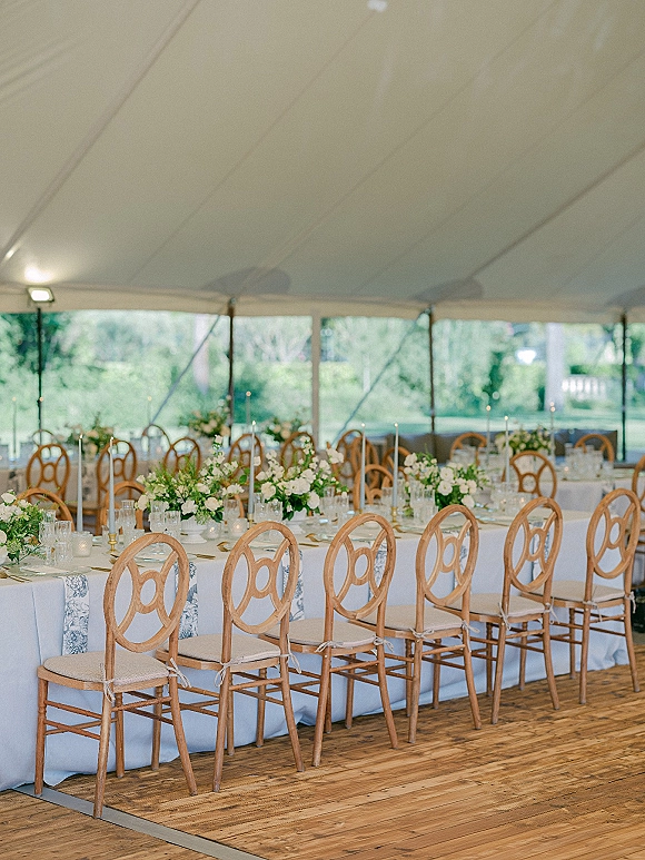 Reception tablescape with long banquet table wedding details, white linens, oval-back wooden chairs, floral centerpieces, and taper candles under a white tent canopy