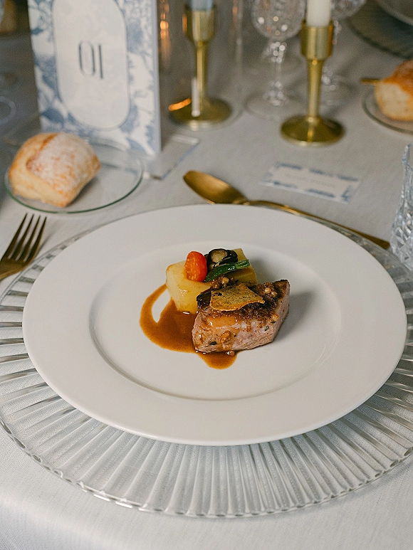 Wedding reception place setting with gold flatware, clear charger and white plate, bread roll, crystal glasses, and blue place card on white linen