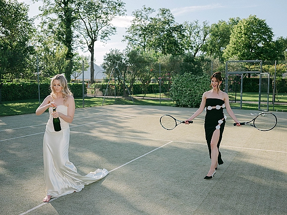 Bridal party portrait of the bride popping champagne beside friends in black gowns holding tennis rackets on an outdoor court with trees behind