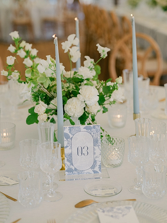 Reception tablescape with a wedding table centerpiece of white florals, blue taper candles, gold flatware and glass goblets in a softly lit room