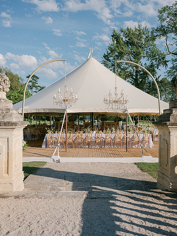 Wedding reception tent with crystal chandeliers over a long white-linen banquet table, set on a lawn near stone gate pillars under blue skies