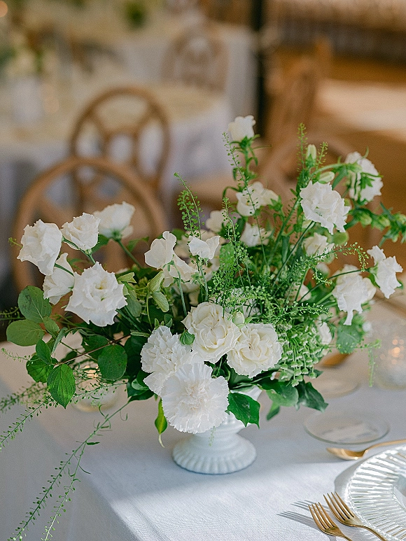 Wedding centerpiece with a white floral centerpiece and greenery in a ceramic compote vase on a table set with gold flatware and glass goblets