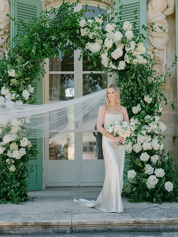 Bridal portrait of a bride holding bouquet in a strapless gown, long veil blowing on steps beneath a hydrangea arch backdrop