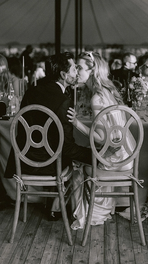 Wedding kiss as bride and groom kissing at sweetheart table, bride in strapless gown with long earrings amid candlelit florals in tent reception