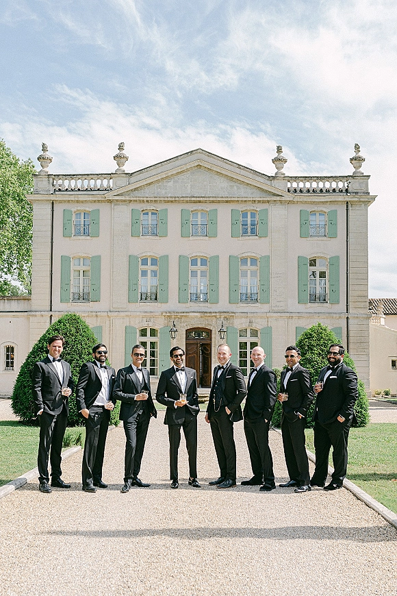 Groomsmen portrait of a black tie lineup in tuxedos and sunglasses holding whiskey glasses on a manor driveway under blue sky