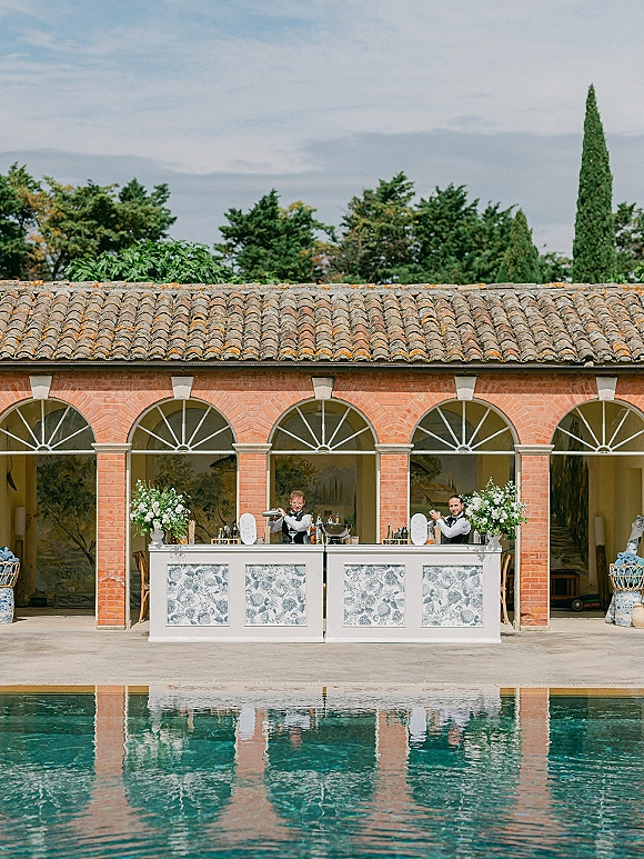 Outdoor wedding bar with a white counter, floral arrangements, bottles and glassware set poolside against brick arches and a tiled roof backdrop