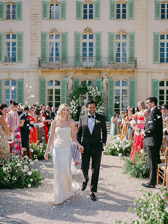 Wedding recessional as bride and groom walk the aisle through a rose petal exit in a sunny stone manor courtyard with guests cheering
