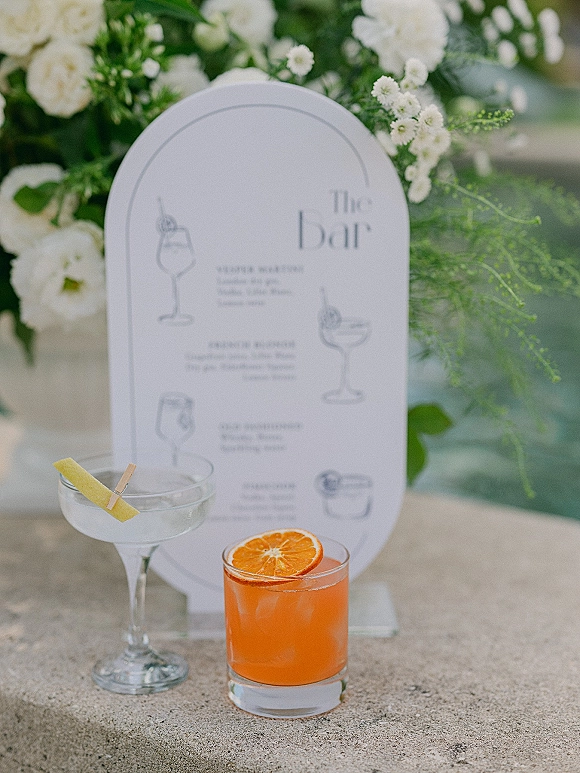 Wedding bar menu and signature cocktail menu on an arched sign with coupe and rocks glasses, citrus garnishes, and florals on stone outdoors