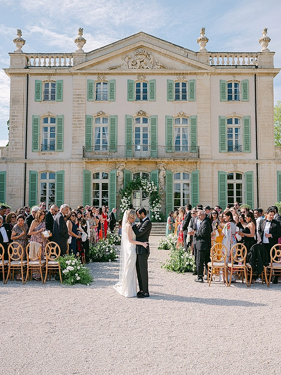 Ceremony kiss moment as bride and groom kiss beneath a white floral arch, veil trailing, with guests in a gravel estate courtyard backdrop