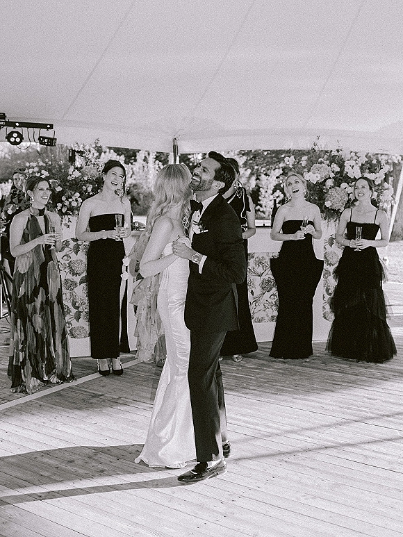 First dance as bride and groom dancing on a wooden floor under an outdoor tent, bridesmaids in black dresses watch near the bar