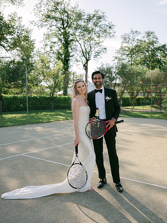 Couple portrait of bride and groom tennis-style, in wedding dress and tuxedo holding rackets on a sunny tennis court by a chain link fence