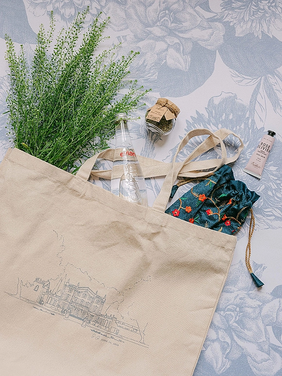 Wedding welcome bag flat lay with a canvas tote, glass water bottle, small jar favor, and hand cream on floral patterned fabric