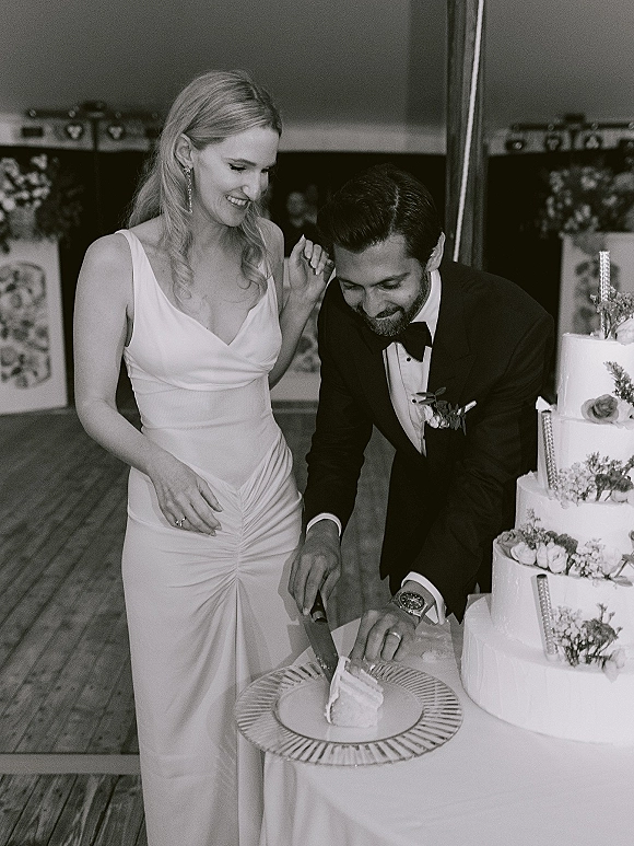 Wedding cake cutting as bride in satin dress and groom in black tuxedo slice a three-tier floral cake on wood floor by folding screens