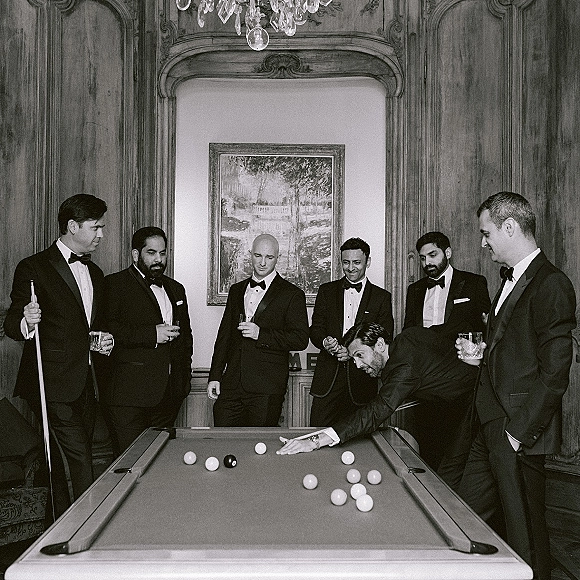 Groomsmen portrait of tuxedoed friends in bow ties holding whiskey glasses around a pool table in a wood-paneled billiards room