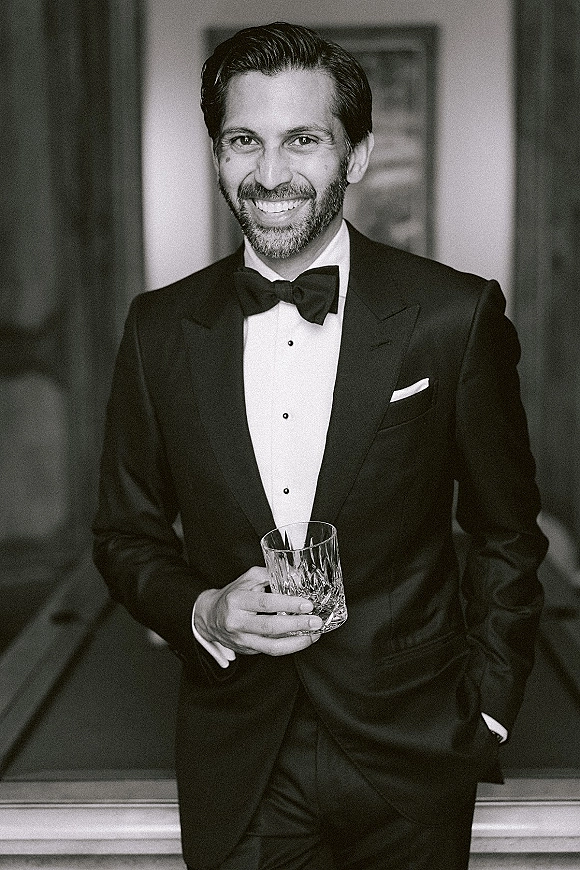 Groom portrait in a black tie tuxedo, bow tie and pocket square, holding a whiskey glass in an indoor room with framed artwork