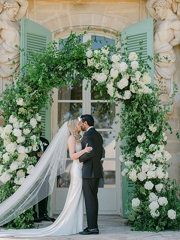 Wedding kiss as couple kisses under arch with white hydrangeas and greenery, bride’s veil flowing before a stone building doorway